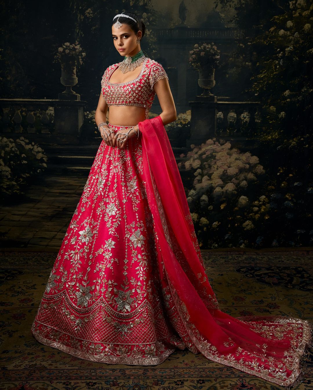 EVARA’s fuchsia raw silk lehenga is elevated by intricate crystal-embellished floral embroidery, balanced with silver, gold, and champagne sequins. The encrusted blouse, with its jeweled back and delicate 